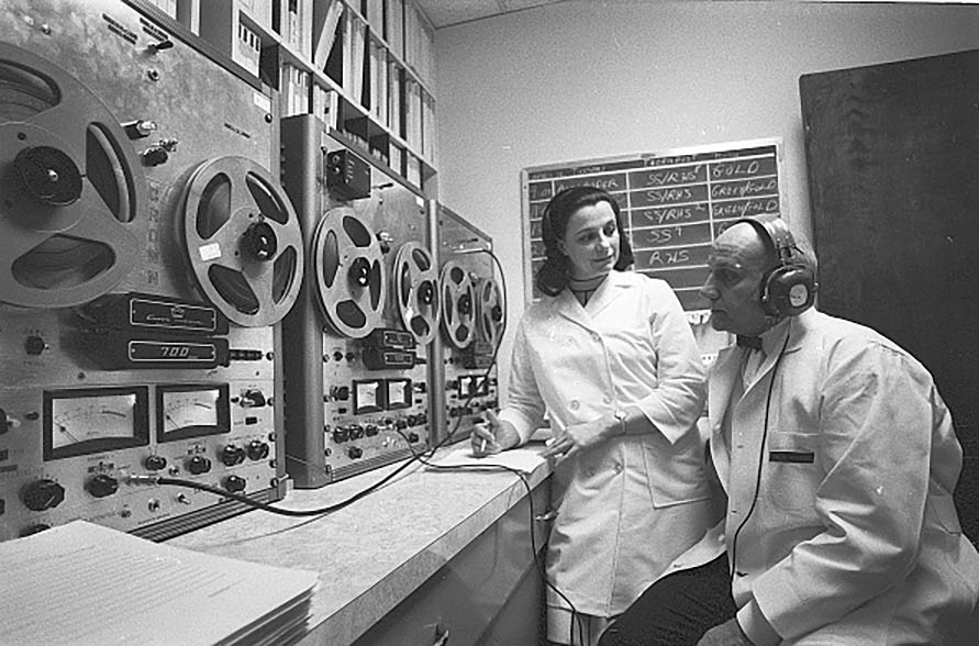 Two individuals in lab coats stand beside a large machine in a laboratory setting.