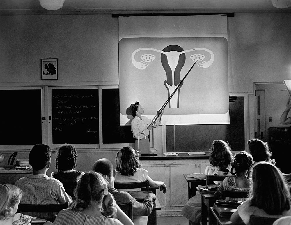 A black and white photo of a classroom with a woman teaching students at desks, engaged in a lesson.
