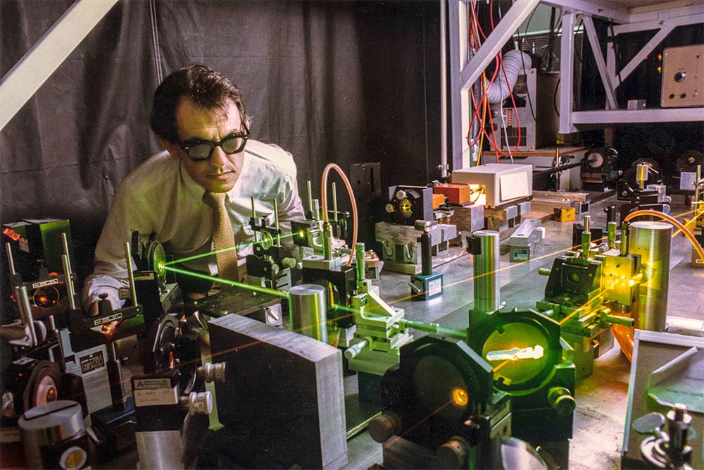 Gerard Mourou, dressed in a white shirt and tie, is engaged in work on a sophisticated machine.