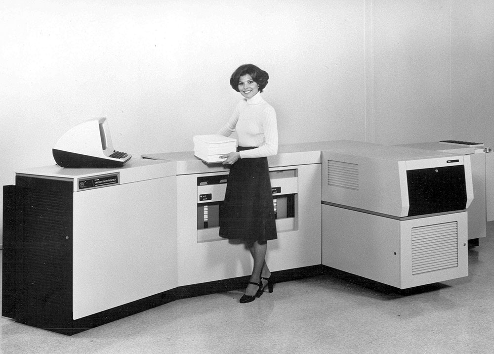 A woman stands in front of a large computer, focused on the screen, with a professional demeanor.