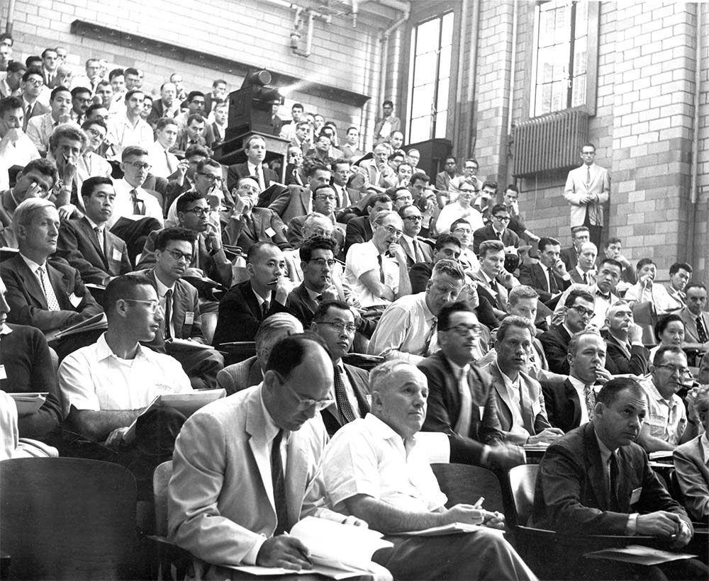 A large audience seated in a spacious auditorium, attentively facing the front stage.