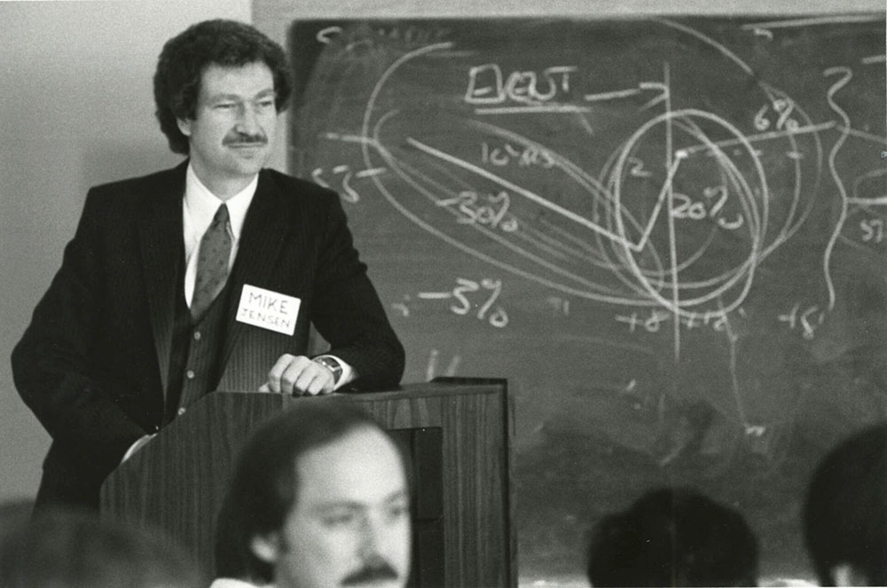 Michael Jensen in a suit and tie stands at a podium in front of a blackboard, addressing an audience.