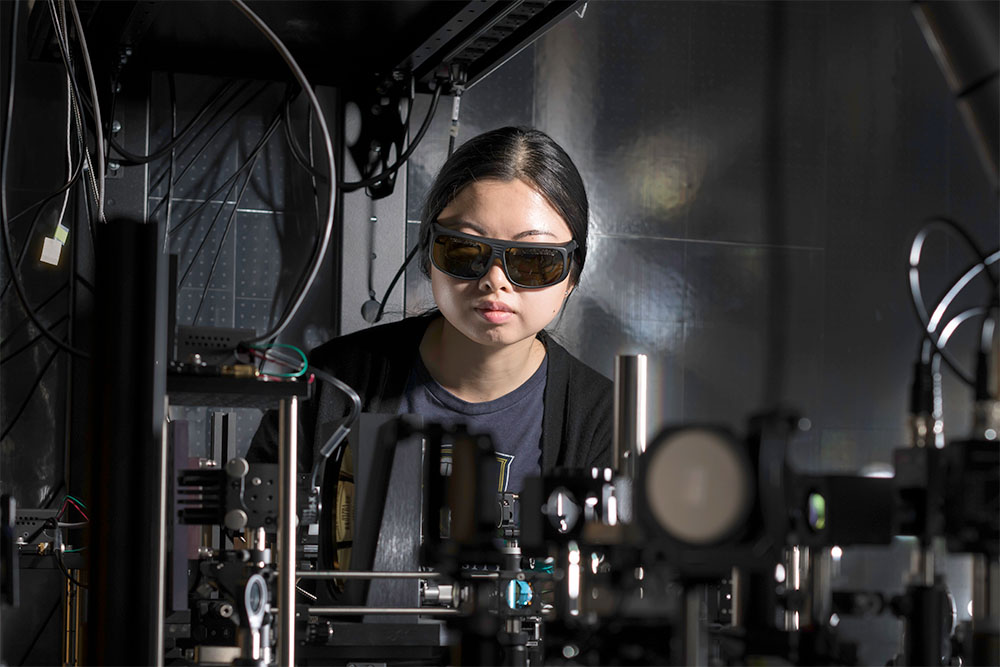 A woman with glasses stands in a lab, focused on operating a complex machine.