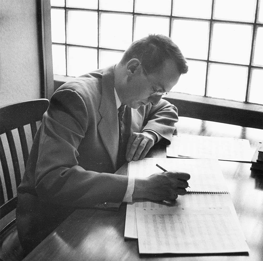 A man in a suit sits at a desk, surrounded by papers and holding a pen, focused on his work.