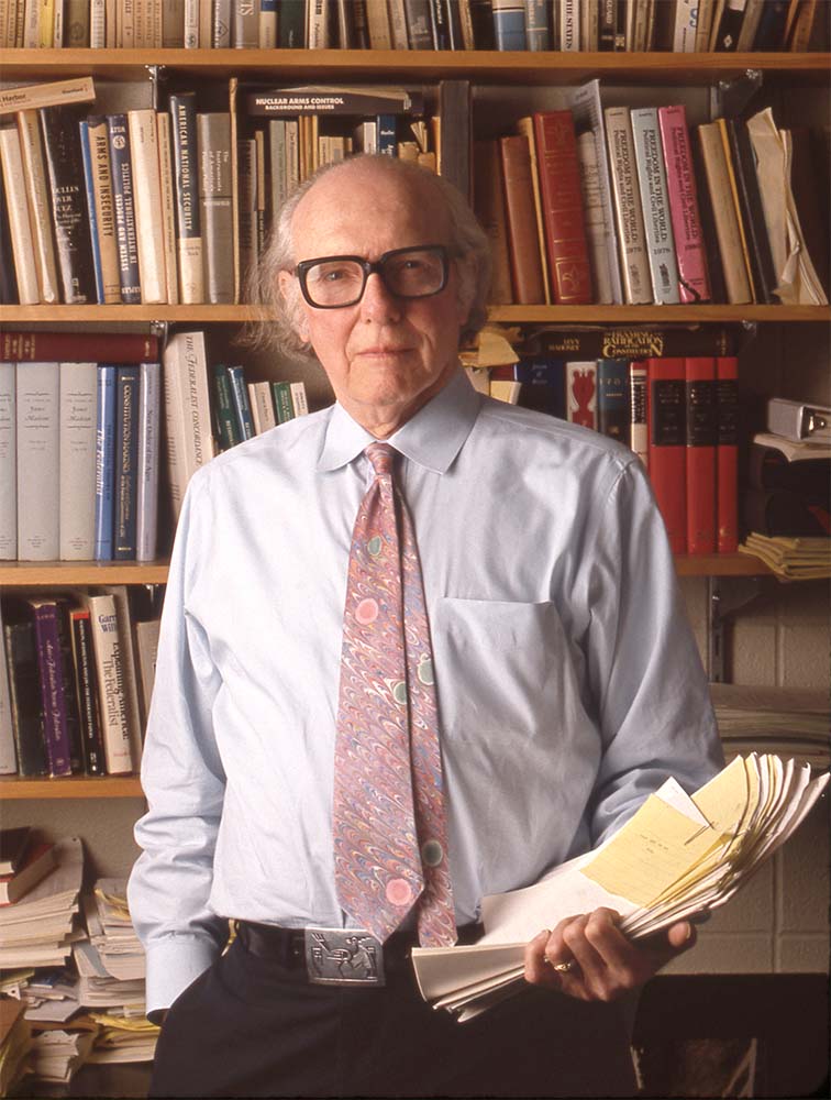 An older man with glasses and a tie, named William Riker, is holding a stack of papers in his hands.