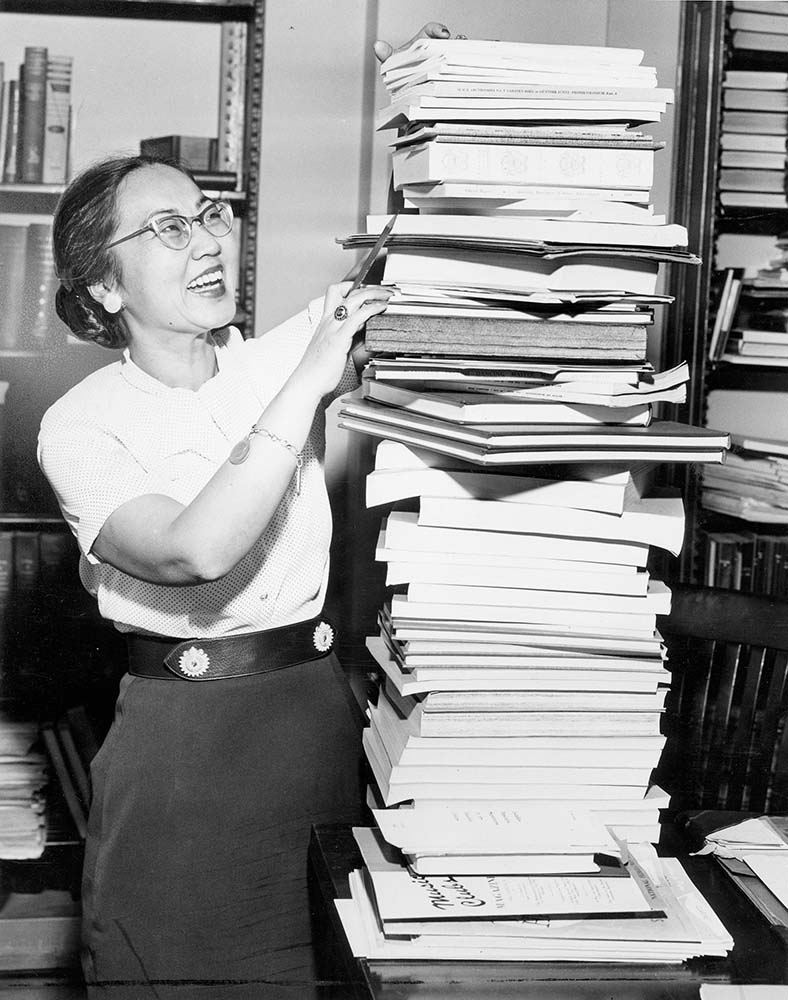 Ruth Watanabe, a woman in glasses and a skirt, holds a stack of books in her arms, smiling at the camera.