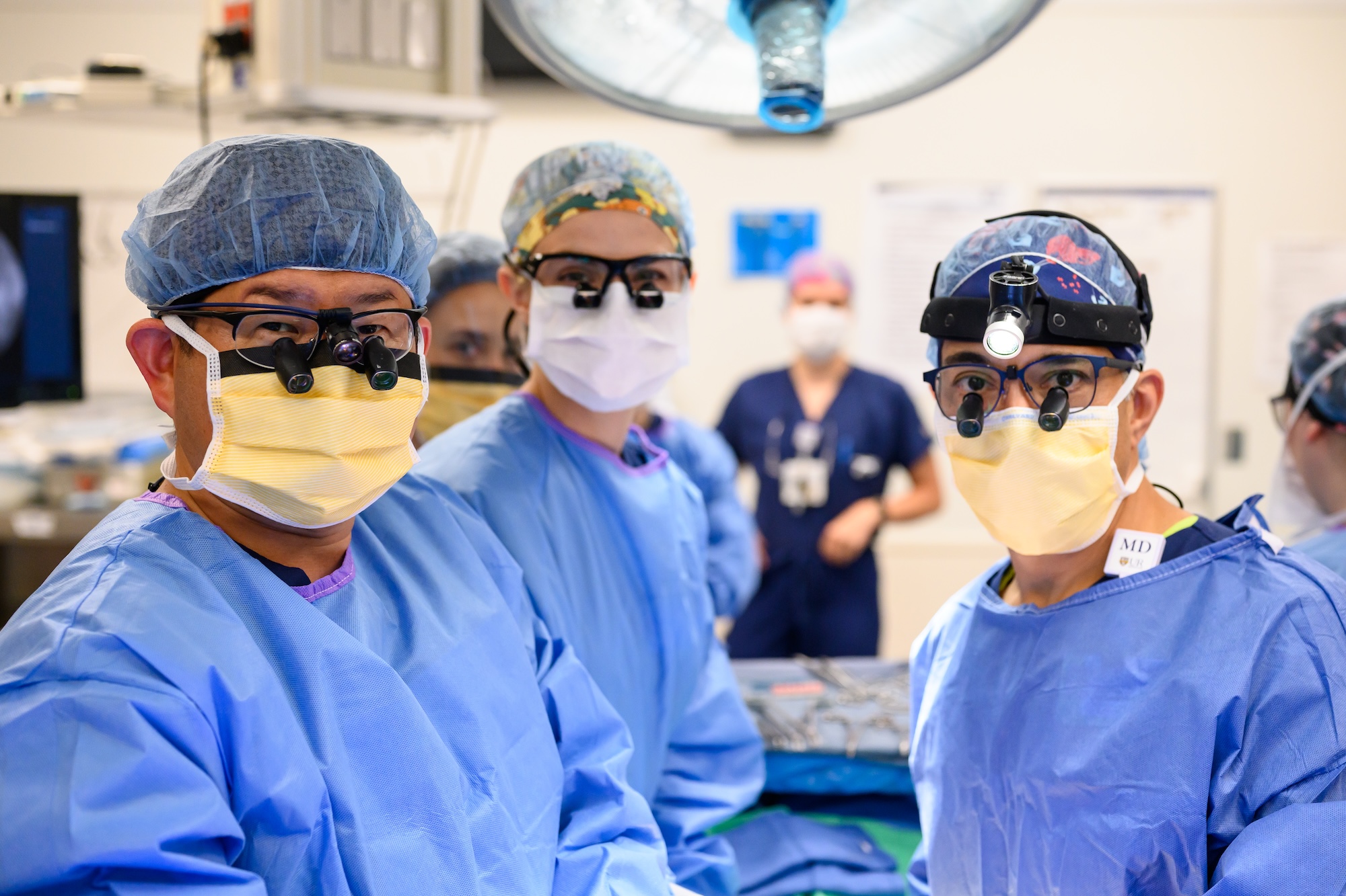 A surgical team in blue gowns and masks works closely Surgeons in blue gowns and masks, wearing headlamps and magnifying loupes, stand together in an operating room during a procedure.