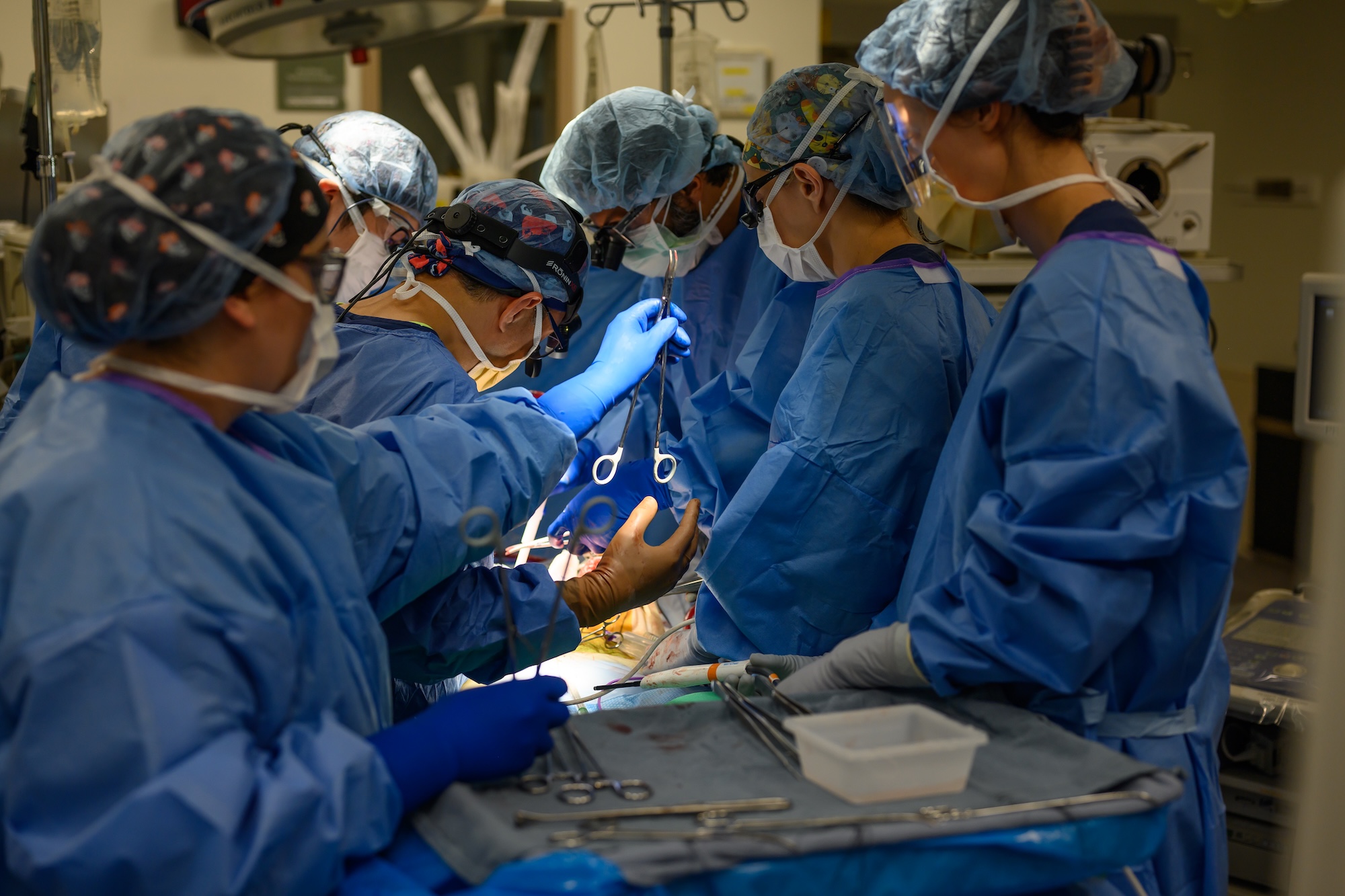 A surgical team in blue gowns and masks works closely together over an operating table, illuminated by bright overhead lights.