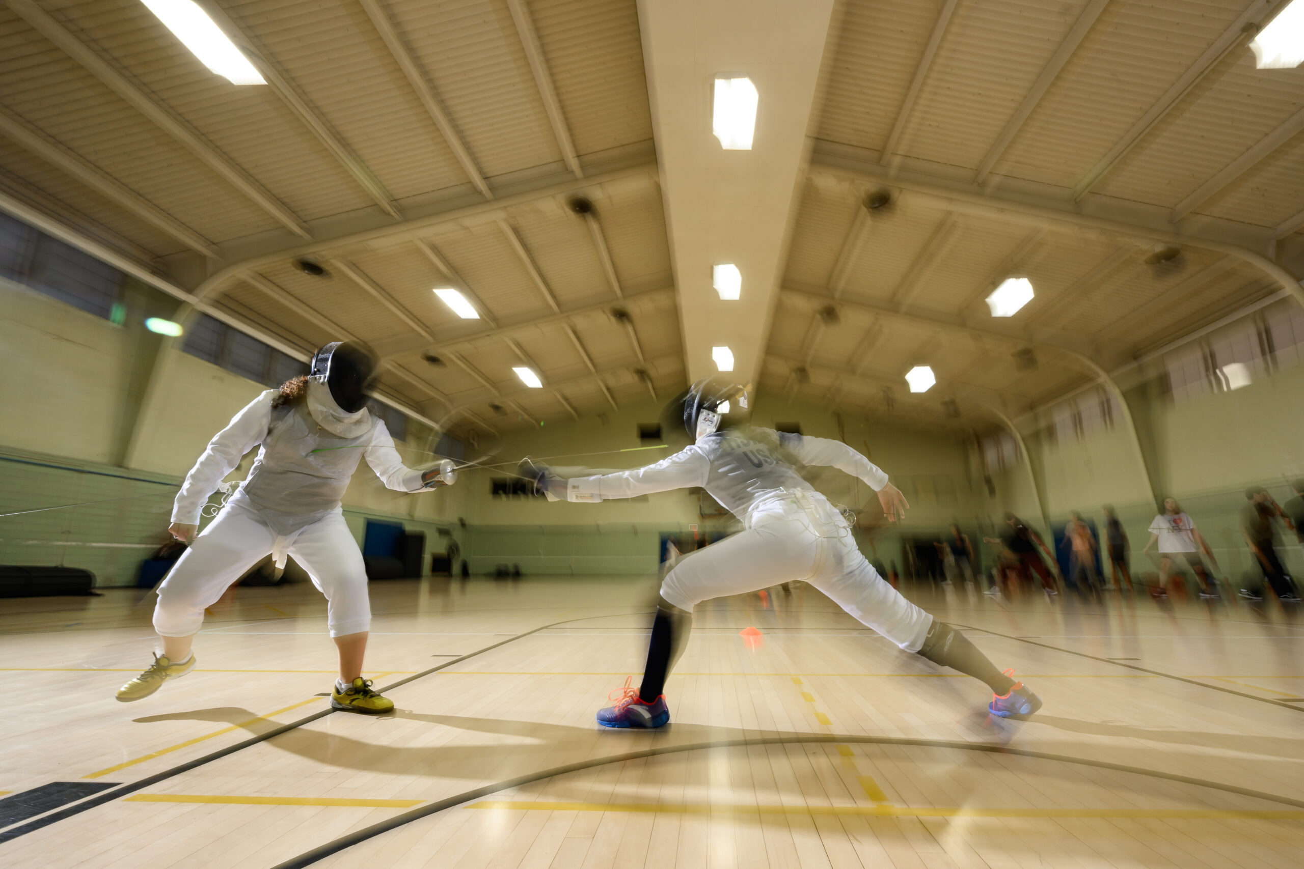 Jackie Hsiao and another URochester fencer in full gear duel in a gym, one lunging forward as their foils meet, captured with motion blur to show speed.
