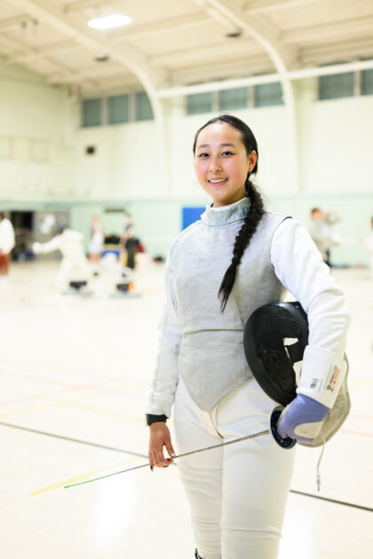 Fencer Jackie Hsiao in full gear smiles while holding her mask and épée in a brightly lit indoor practice gym.