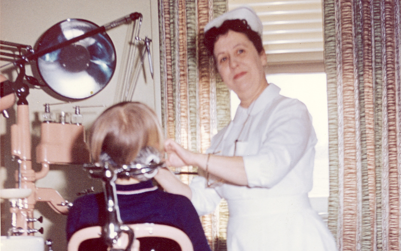 Dental hygienist in uniform treats a child in a vintage dental chair, part of an early school dental care program.