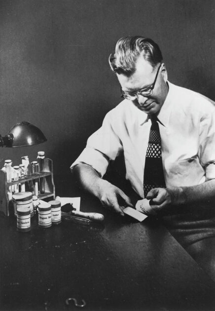 A man in glasses and a tie sits at a desk with a bottle of pills, referencing Carlson's 1938 experiment.