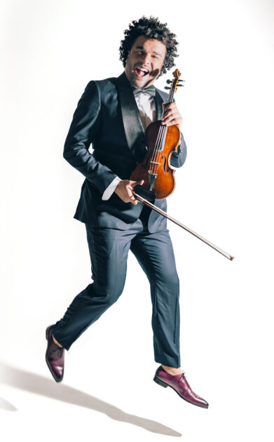 Violinist Curtis Stewart leaps joyfully while dressed in a tuxedo and holding his violin and bow.