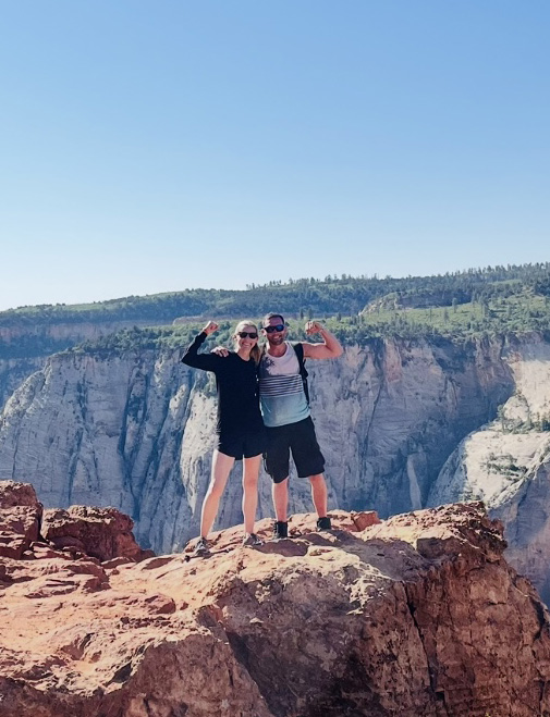 Two hikers stand on a rocky cliff edge with arms raised, smiling against a backdrop of steep canyon walls and a clear blue sky.