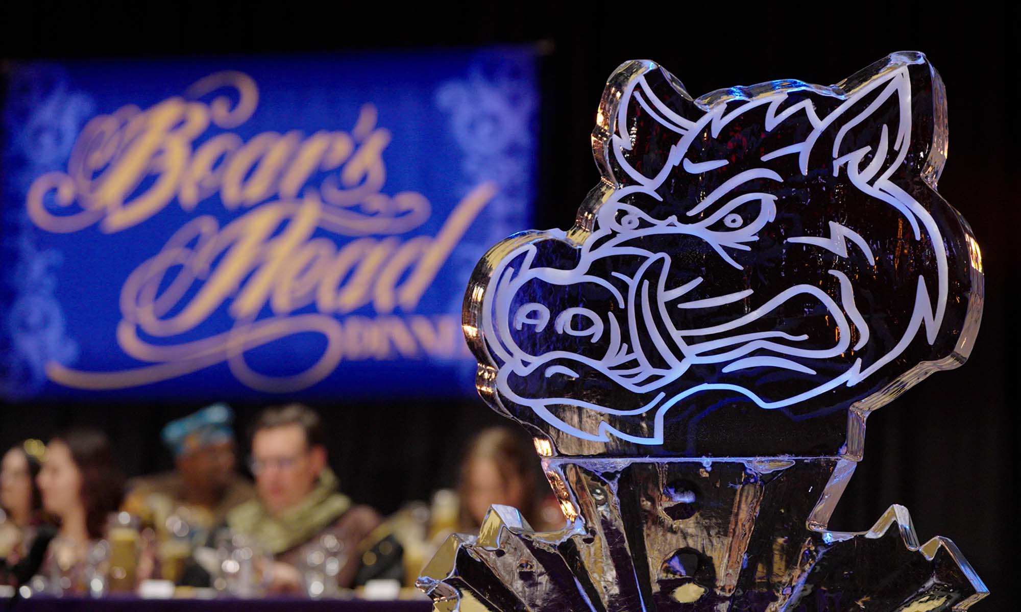 Close up of an ice sculpture of a boar's head with a screen in the background that reads "Boar's Head Dinner" and faculty in medieval garb at a high table.