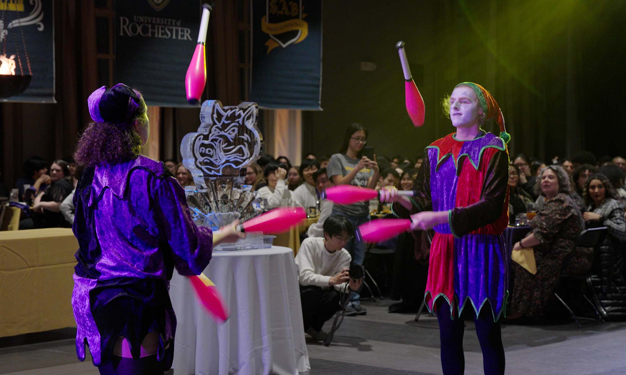 University of Rochester undergraduates in medieval garb juggling in front of an ice statue of a boar's head during the 2024 Boar's Head Dinner.