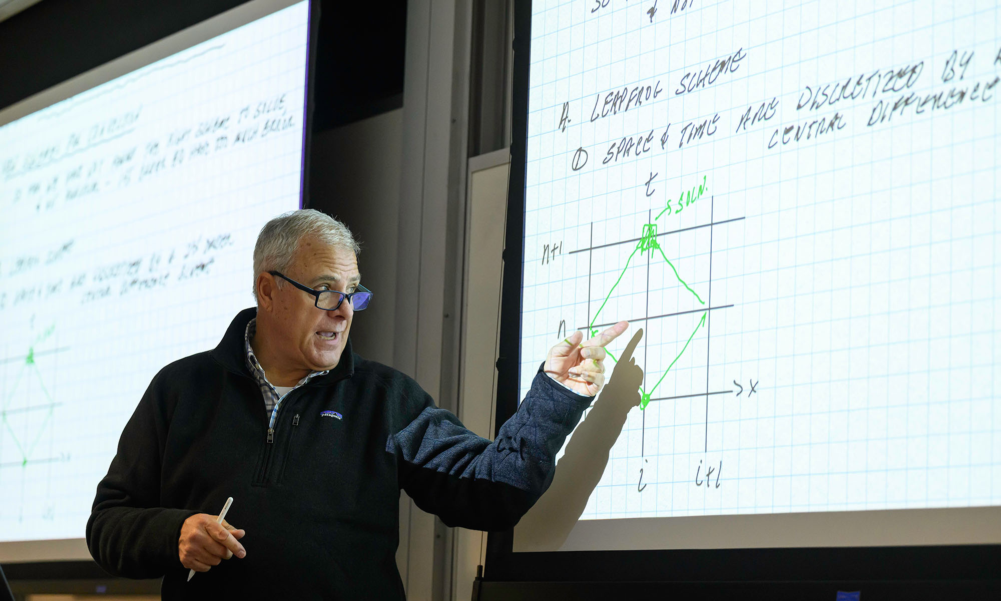 David Foster in a classroom teaching and gesturing at a screen full of graphs and equations.
