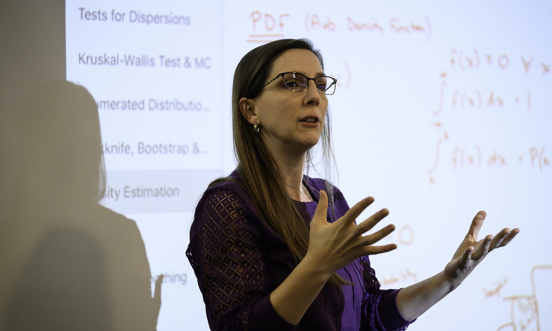 Katherine Grzesik in a classroom teaching and gesturing at a screen with equations.