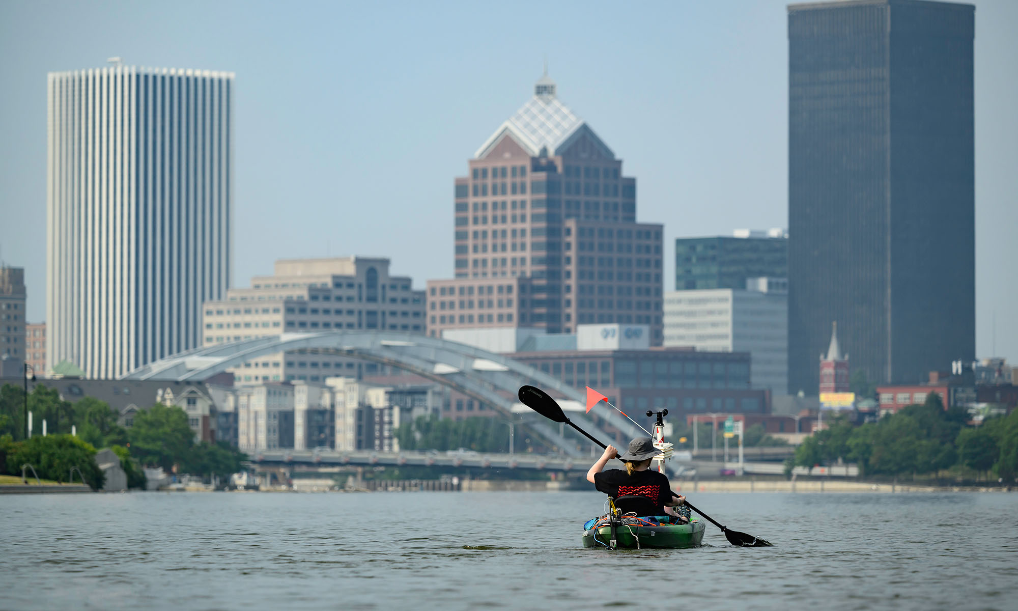 A kayaker seen from behind on the Genesee River with the city of Rochester skyline.