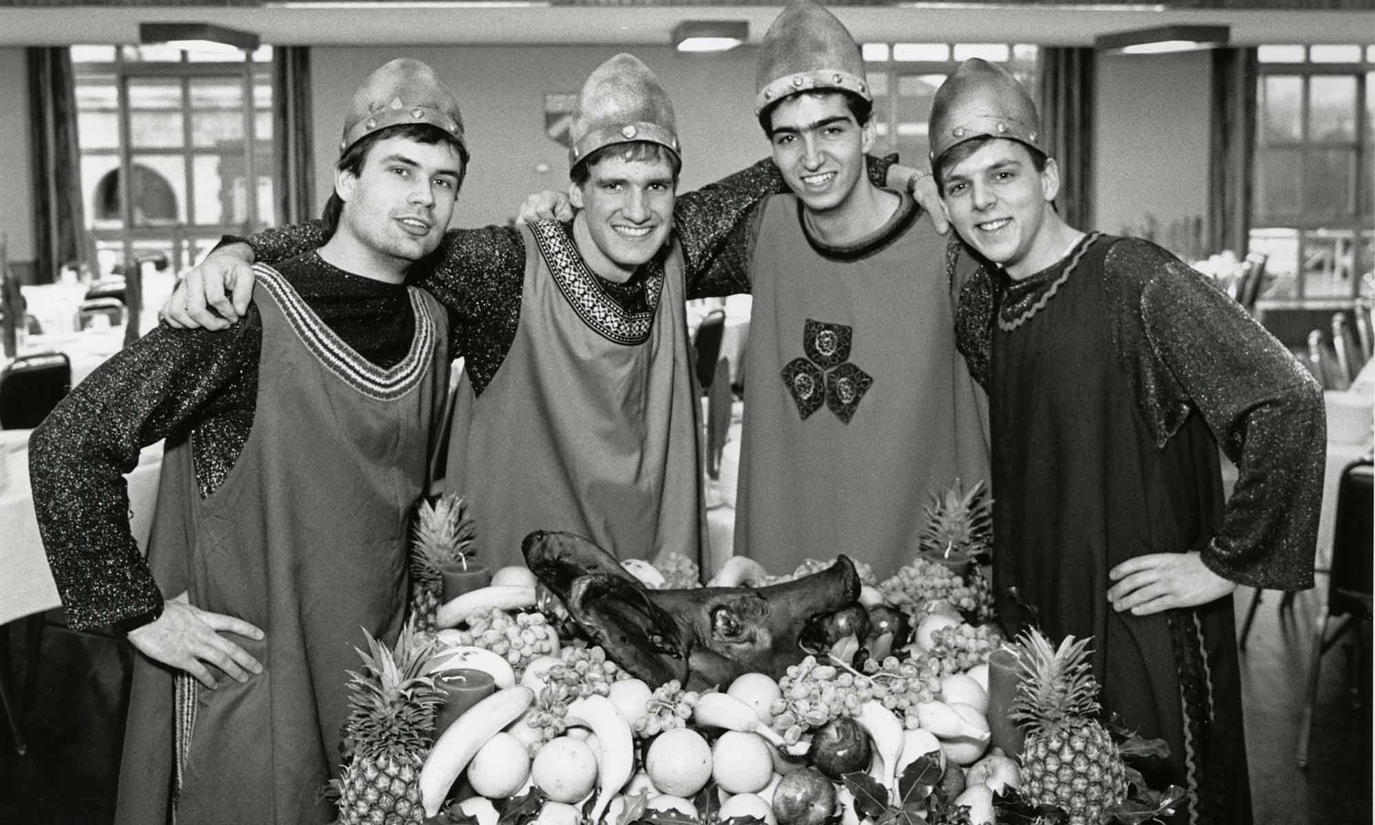 Four male University of Rochester undergraduates in medieval garb smile at the camera while posed around a table with a boar's head and trimmings.