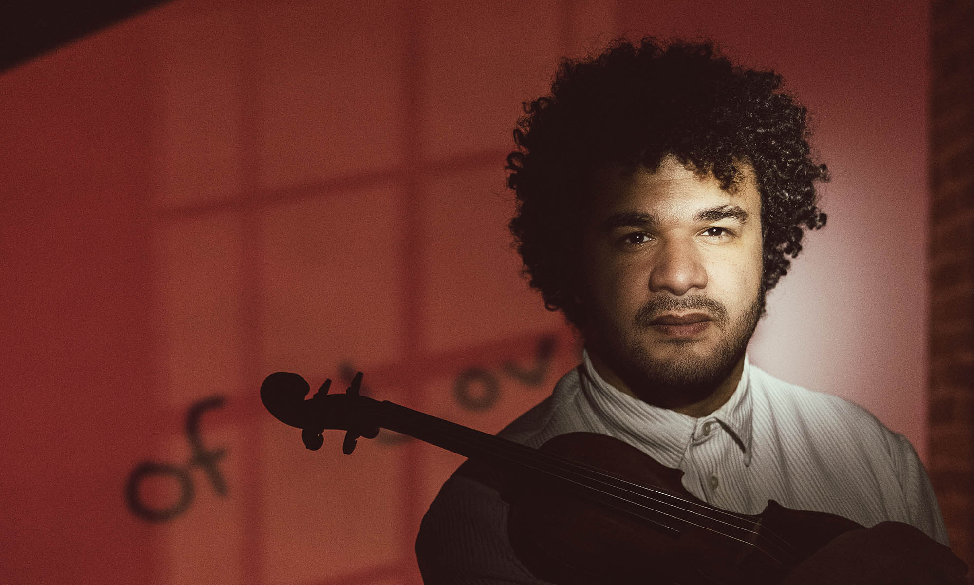 Portrait of Curtis Stewart holding a violin, lit dramatically against a reddish background with soft shadowed text.
