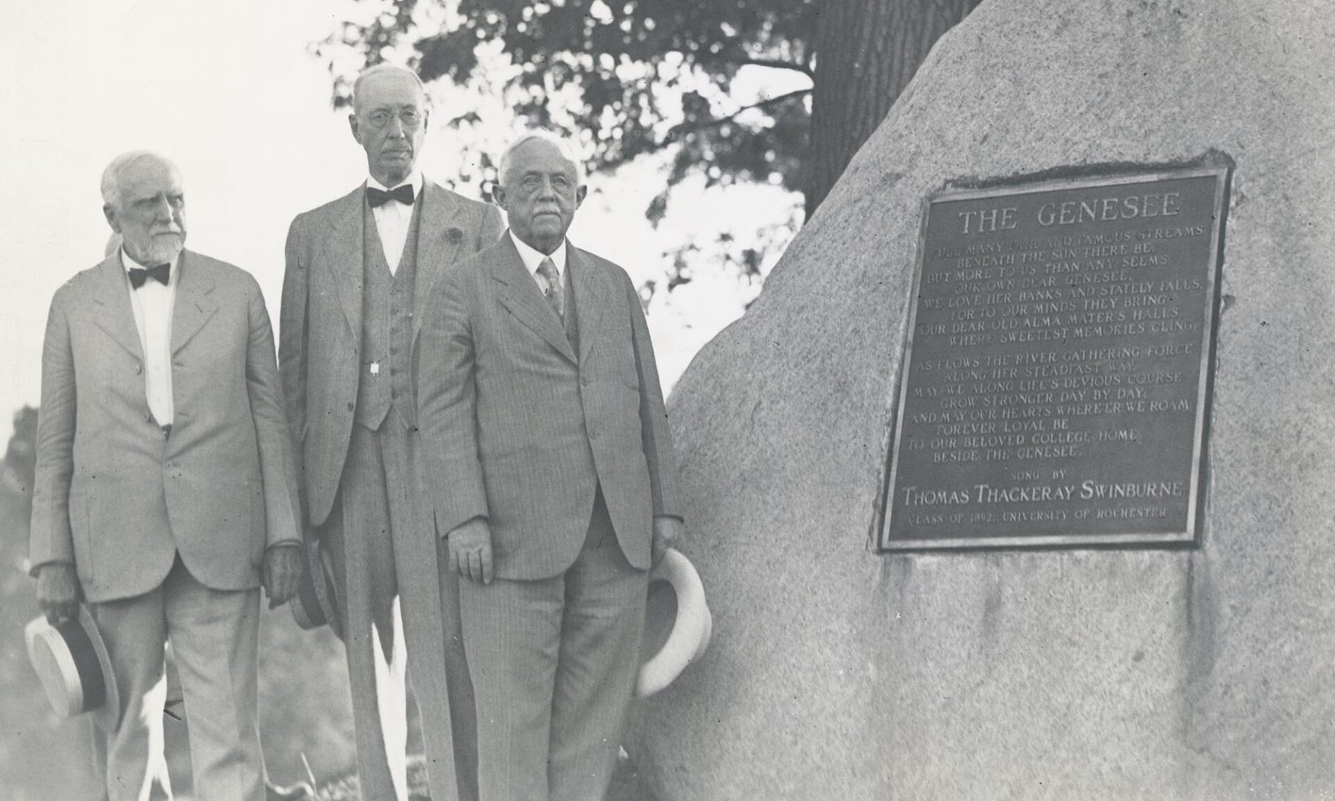 Three men in suits stand beside a stone monument engraved with the University of Rochester alma mater “The Genesee.”