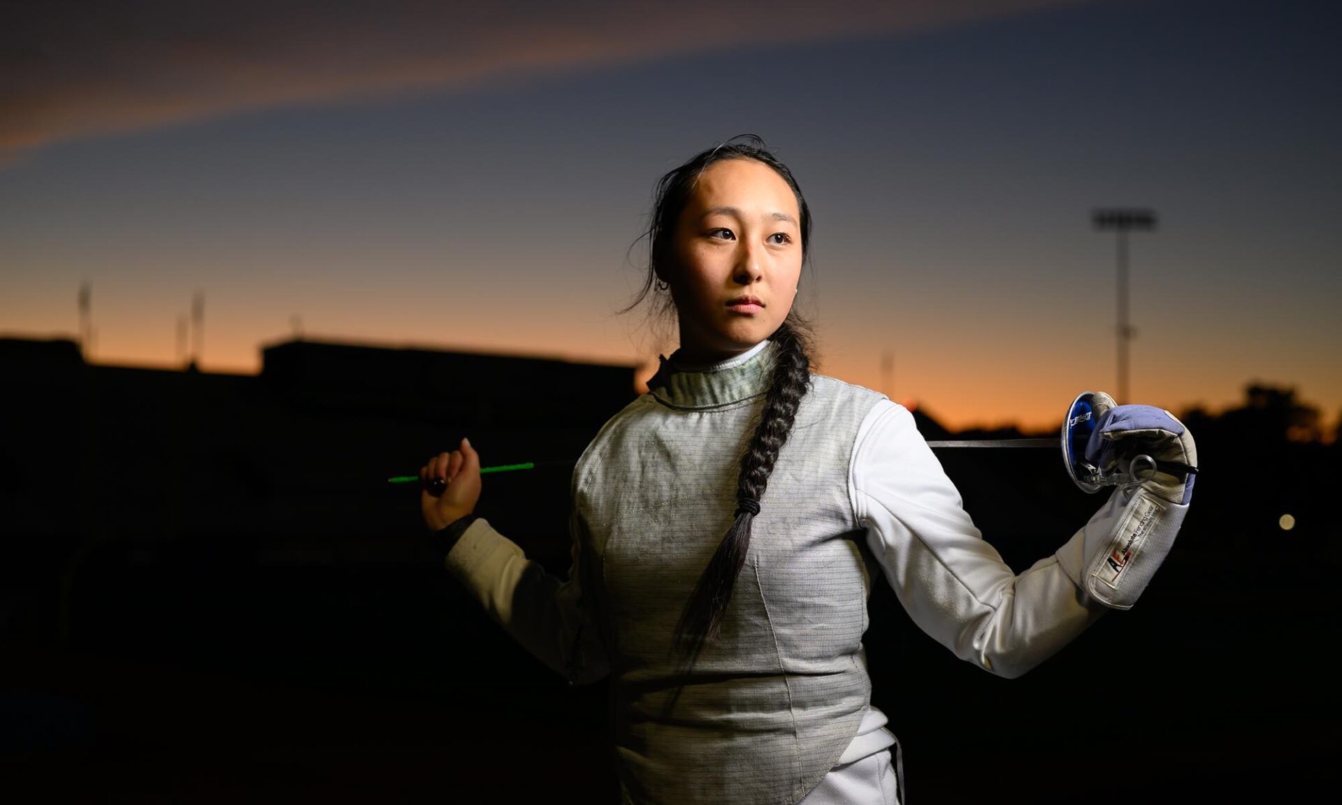 Fencer Jackie Hsiao poses confidently at sunset, holding her foil across her shoulders and wearing full fencing gear.