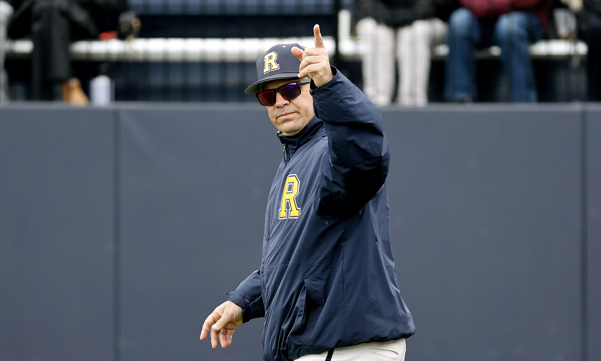 University of Rochester baseball coach Joe Reina in team gear points toward the sky during a game.