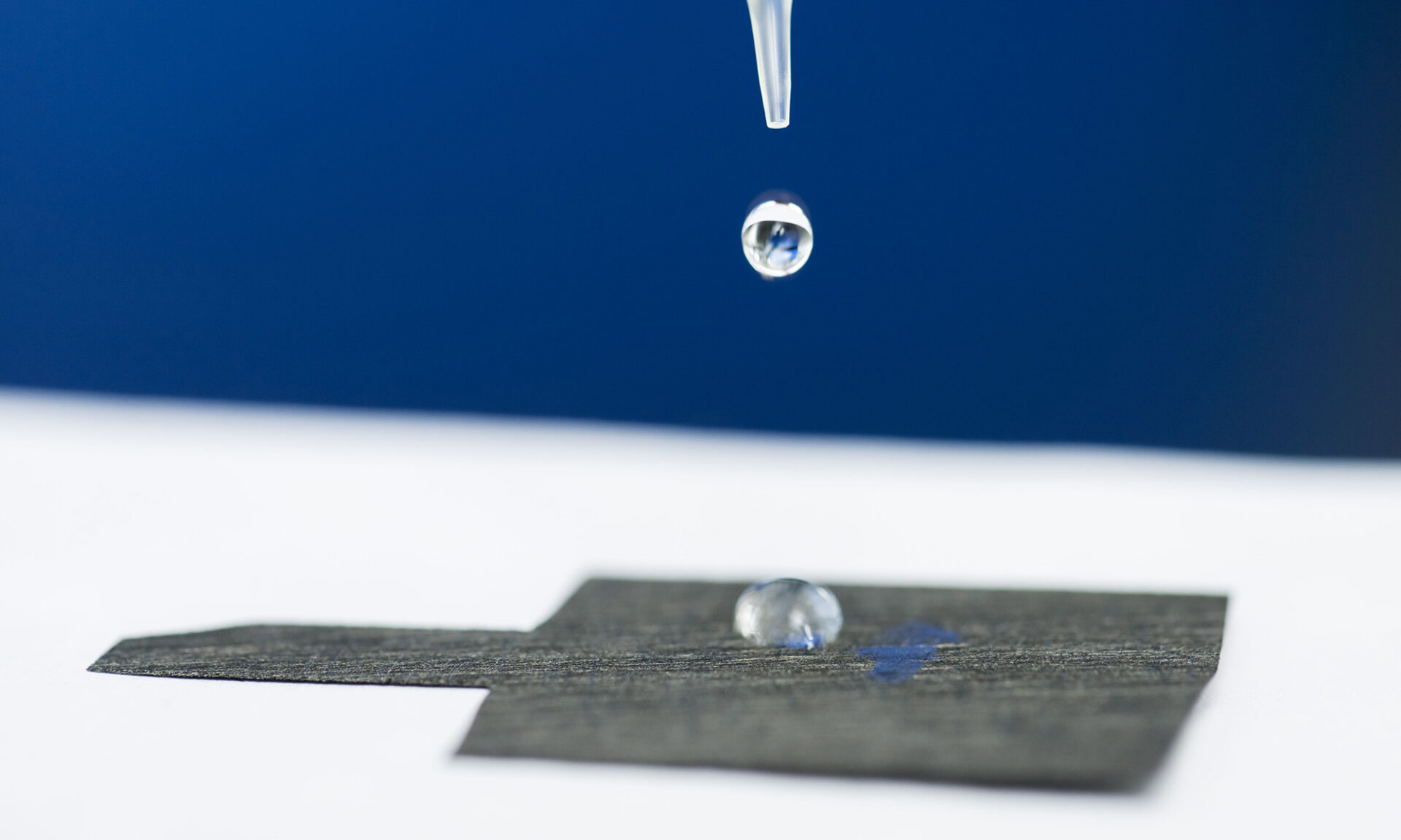 A pipette releases a drop of water onto a piece of water-attracting carbon paper with a water droplet already adhered to it.