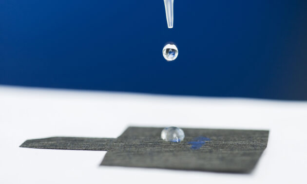 A pipette releases a drop of water onto a piece of water-attracting carbon paper with a water droplet already adhered to it.