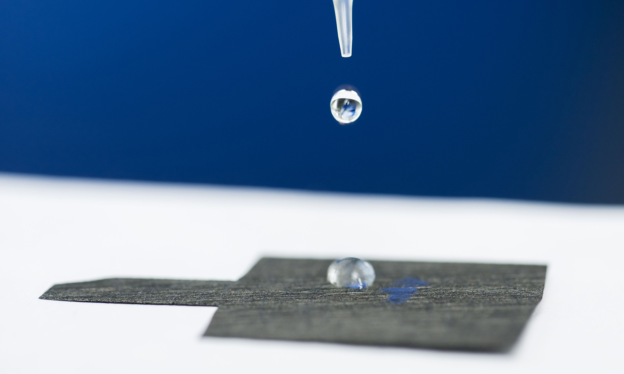 A pipette releases a drop of water onto a piece of water-attracting carbon paper with a water droplet already adhered to it.