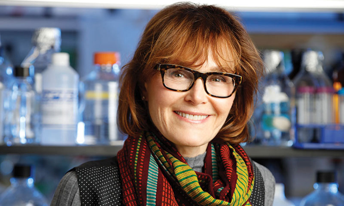 A woman with glasses and a scarf stands in front of a lab at the University of Rochester, focusing on RNA biology research.