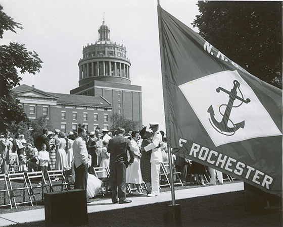 Black and white archival photo of a University of Rochester NROTC flag in the foreground, NROTC cadets and families in the background, with Rush Rhees Library.