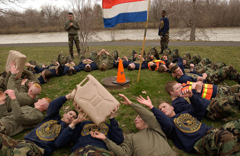 University of Rochester NROTC students lay on the ground outside by the Genesee River in a circle and pass a heavy container to each other as part of a training exercise.
