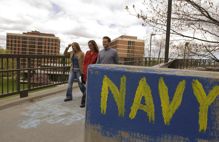 Three URochester students walking on campus by a concrete post painted blue with the word 