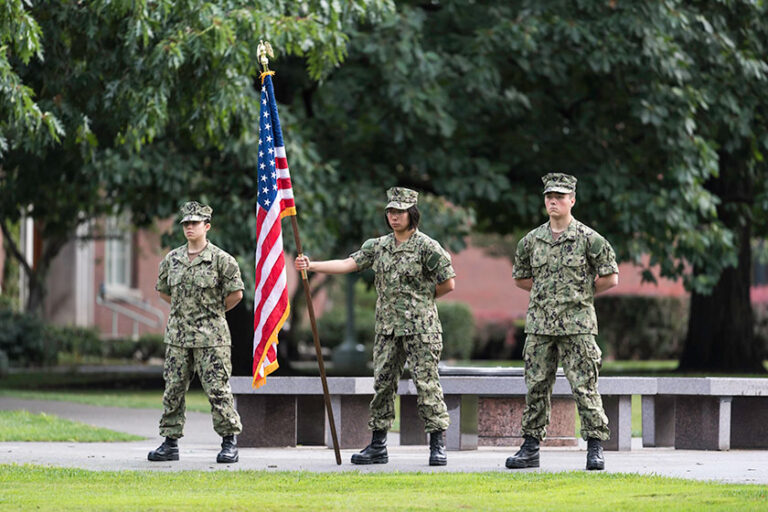 Three NROTC midshipmen in uniform and holding a flag during a 9/11 vigil on campus.