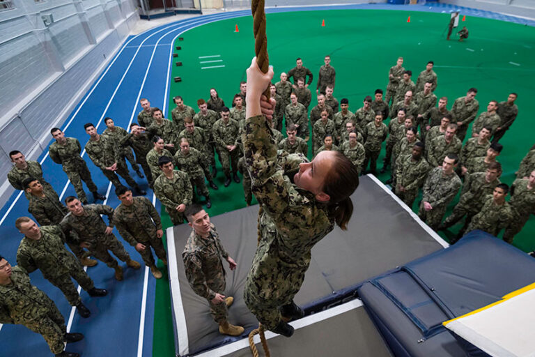 An NROTC cadet climbs a rope while fellow midshipmen look on.