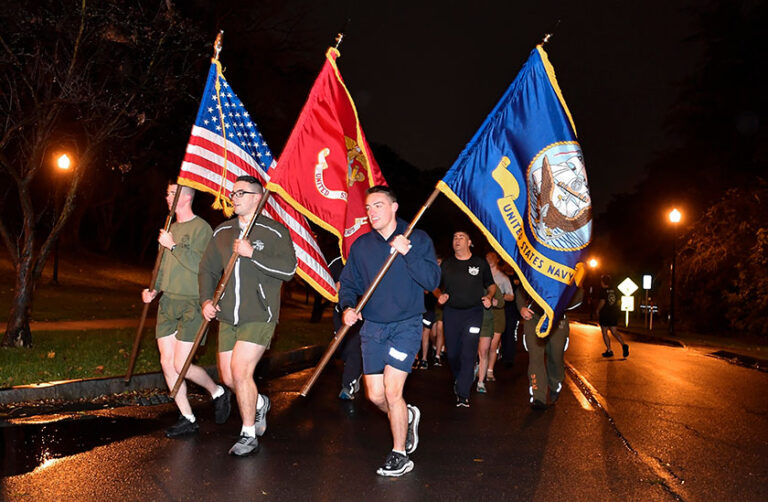 Veterans carrying flags for the US, Marines, and Navy lead a group of runners in the early morning hours.