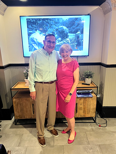 George Ward ’65 and his wife, Peggy, smile at the camera while posed in front of an archival picture from 1965 on the screen behind them.