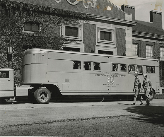 Black and white archival photo of an NROTC bus on the URochester campus with a pair of cadets walking nearby.