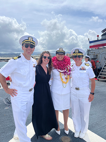 Four people (three in Navy whites) at a naval base pose together for a photo.