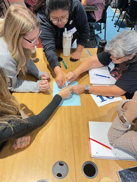 Near overhead view of several math educators from New York State rural schools huddled around a table working on a team-building exercise. 