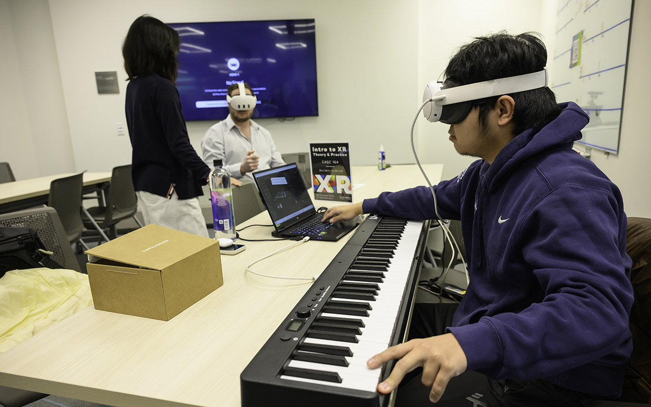 A student wearing a VR headset plays a digital keyboard at a table while classmates test VR tools in a classroom.