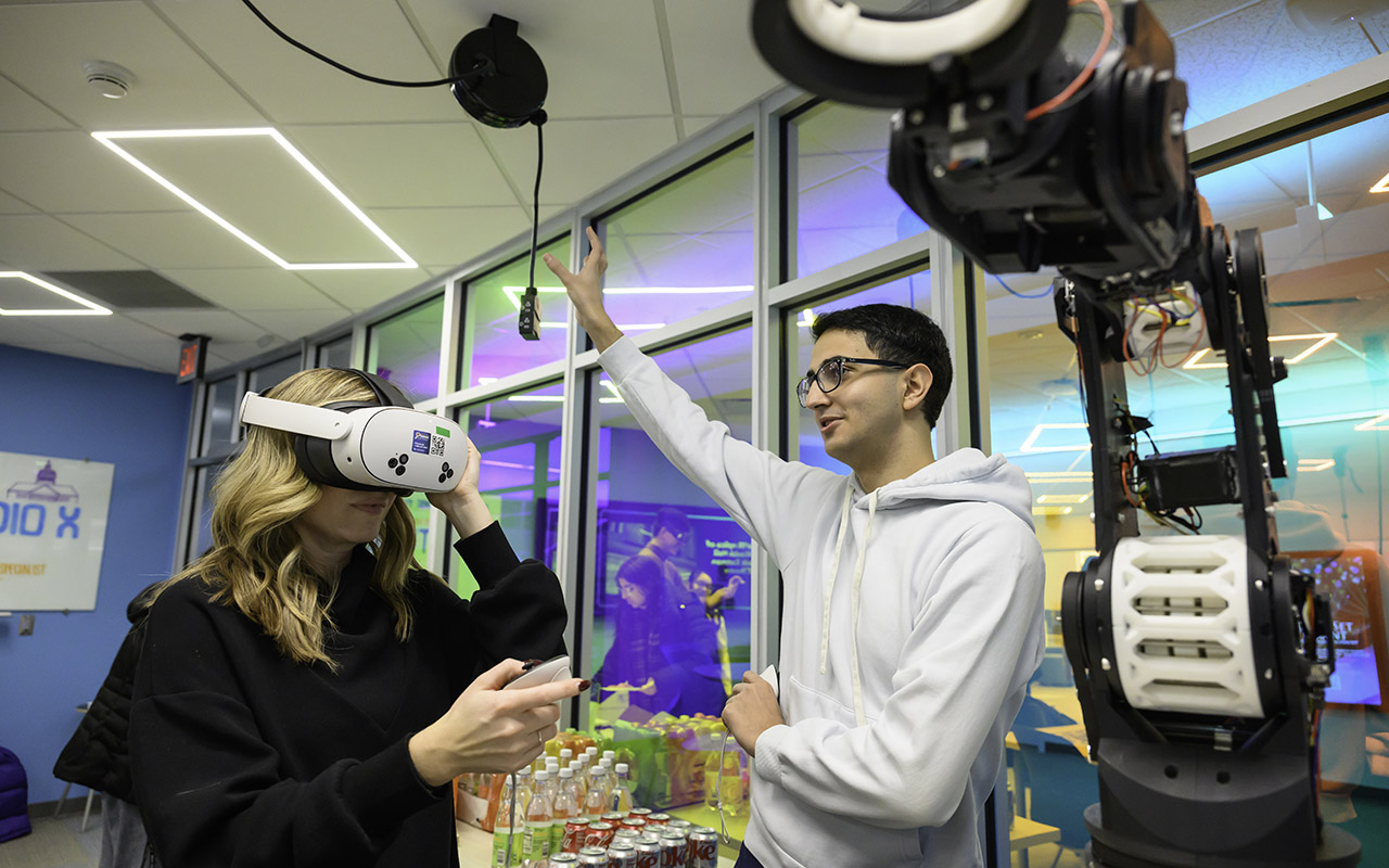 A student wears a VR headset and holds a controller while another gestures nearby, with a robotic arm and colorful lab space behind them.