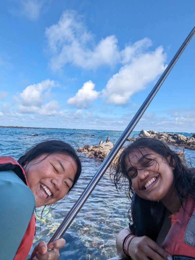 Christine Bresnehan and a friend in life jackets lean toward the camera on clear blue water, with rocky islets and small birds visible behind them.