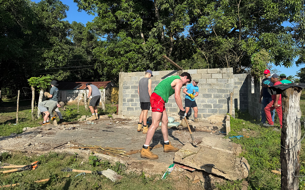 Students and local workers clear rubble and break up concrete at a construction site, surrounded by trees and partially built block walls.