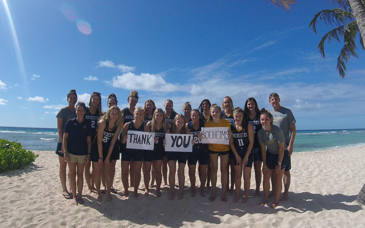 A large group of students stands barefoot on a sunny beach holding a “Thank You” sign, with palm trees, blue sky, and ocean behind them.