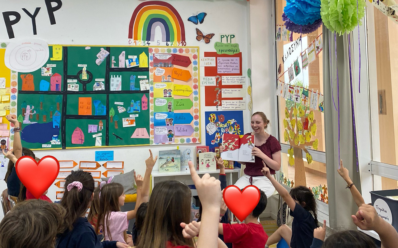 Katie Hurwitz reads to a lively classroom of young children, who sit on the floor with hands raised amid colorful artwork and posters.