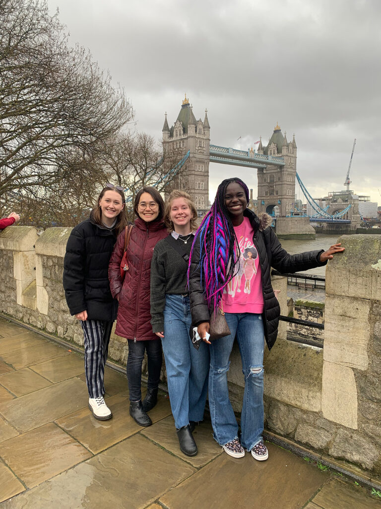 Four friends pose in front of London’s Tower Bridge on a cloudy day, smiling as they stand along a stone walkway.