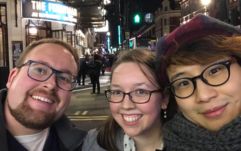 Three friends smile for a nighttime selfie on a busy London street, with bright theater signs and crowds behind them.